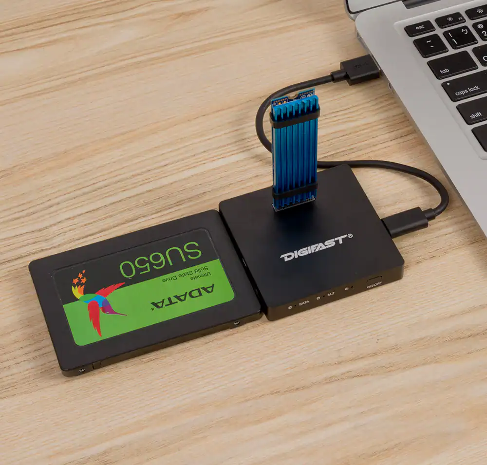 A laptop on a wooden surface connects via USB to a Digifast DX3 Docking Base Ultra High Speed, which holds an ADATA SU650 SATA SSD and an M.2 SSD with a blue heatsink.